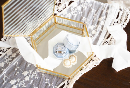 Hexagonal glass jewelry box with silver rings and pearl earrings on lace, white ribbon accent, perfect Louisiana wedding inspo.