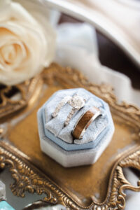 Diamond engagement ring and wooden wedding band on blue velvet box, gold tray, and white rose—Louisiana wedding inspiration.