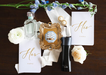 Wedding flat lay with vow books, perfume, cologne, earrings, rings on a gold tray, and flowers on dark wood.