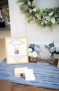 Gulf South-inspired wedding table with card and guest book signs, blue flowers, wooden box on blue cloth, and green wreath backdrop.