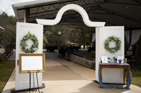 White doors with wreaths welcome guests to a Gulf South wedding venue, with a gold sign left and guest gift table right, under metal roof.