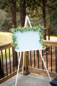 Wedding welcome sign with green leaves on easel, reading: Welcome to our wedding. Pick a seat, not a side. Outdoor setting.