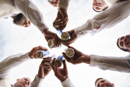 Six men in white shirts toast canned drinks in a circle, celebrating together under the open sky—ideal for Gulf South weddings.