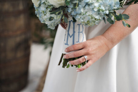 Bride’s hand with diamond ring holds white and blue bouquet wrapped in MB cloth; wooden barrel blurred in background.