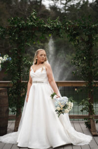 Bride in white dress with bouquet stands on wooden deck by leafy arch and fountain at romantic Louisiana wedding ceremony.