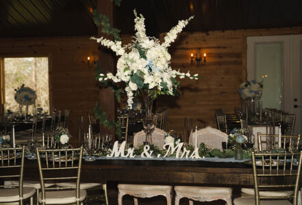 Romantic Louisiana wedding table with white floral centerpiece, gold chairs, Mr. & Mrs. sign in rustic venue with soft lighting.