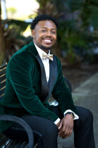Groom portrait in emerald velvet tuxedo jacket with black lapels and gold bow tie, seated outdoors with greenery background