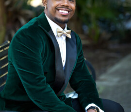 Groom portrait in emerald velvet tuxedo jacket with black lapels and gold bow tie, seated outdoors with greenery background