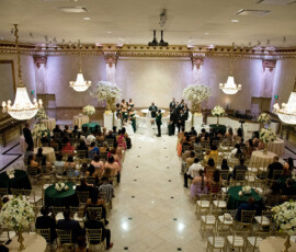 Wide overhead view of grand ballroom wedding ceremony with chandeliers, round guest tables, and floral arrangements framing the aisle