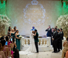 Couple shares first kiss during elegant ballroom wedding ceremony with green uplighting and floral arrangements, monogram reading Tremika and Quentin