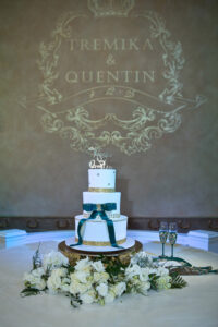 Four-tier white wedding cake with emerald ribbon and gold detailing displayed beneath custom Tremika and Quentin monogram projection