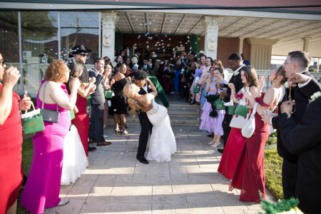 Bride and groom dip kiss outside venue surrounded by guests blowing bubbles during joyful wedding exit celebration