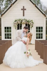 Groom in beige suit dips bride in white gown outside flower-adorned Louisiana chapel during intimate wedding ceremony.