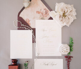 Elegant flat lay of wedding invites, pearl necklace, vintage brooch, rings, and flowers on light gray background.