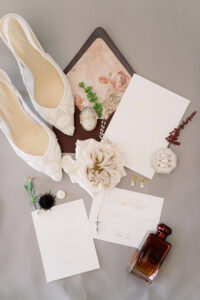 Wedding flat lay: lace shoes, floral invites, white rose, jewelry, perfume bottle, ring box, and small flowers on gray surface.