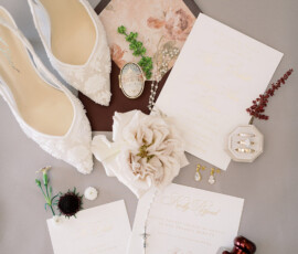 Wedding flat lay: lace shoes, floral invites, white rose, jewelry, perfume bottle, ring box, and small flowers on gray surface.
