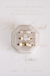 Three gold rings, including a diamond engagement ring, on a gray velvet box with an elegant invitation blurred behind.