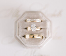 Three gold rings, including a diamond engagement ring, on a gray velvet box with an elegant invitation blurred behind.