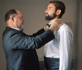 Two bearded men in formalwear indoors as one adjusts the other’s bow tie, preparing for a New Orleans wedding.