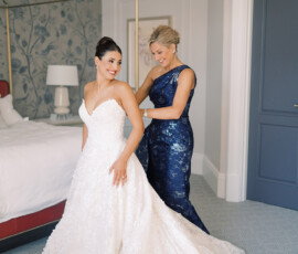 Bride in white gown smiles as woman in navy dress helps her in a bright, elegant bedroom before New Orleans wedding.