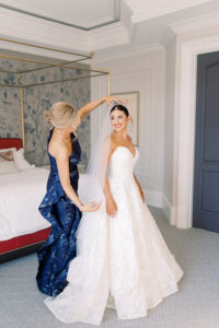 A woman in a navy gown fixes a smiling bride’s veil in a bright bedroom before their New Orleans wedding ceremony.