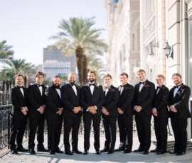Ten men in black tuxedos line up on a city balcony with palm trees, smiling at the camera—New Orleans wedding inspiration.