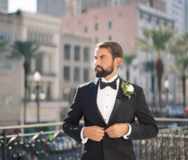 Man in black tuxedo buttons jacket on sunny balcony with city buildings and palm trees, perfect Louisiana wedding inspiration.