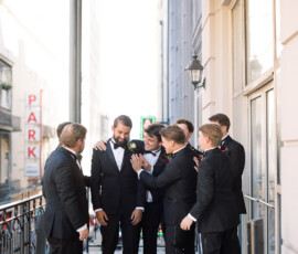 Six men in tuxedos smile and talk on a balcony at a New Orleans wedding, supporting one man in the center.