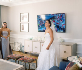 Bride in white dress and veil smiles with gray-dressed bridesmaids in living room, TV showing sports, Louisiana wedding.