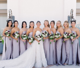 Bride in white gown with eight bridesmaids in lavender dresses, holding bouquets and smiling before a white wall with plants.