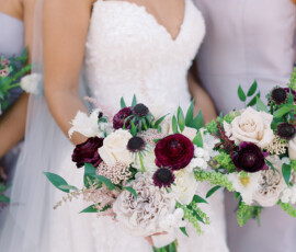 Bride in a white lace gown and veil smiles with bridesmaids in strapless dresses, holding red, white, and blush bouquets.
