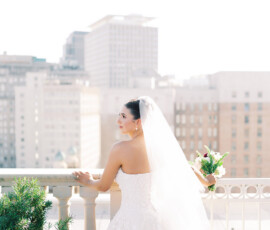 Bride in white dress and veil holds bouquet on balcony, gazing at city skyline on a sunny Gulf South wedding day.