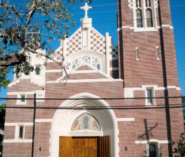 Large brick church with white trim, arched doors, stained glass, and tall bell tower under a clear blue sky.