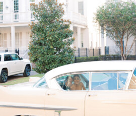 Smiling women sit in a vintage cream car, one waving, on a white house-lined street at a joyful New Orleans wedding.