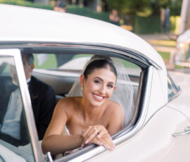 Bride with veil smiles from classic white car window; trees and building in background at Louisiana wedding.