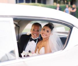 Bride in white dress and veil smiles beside tuxedoed man in classic white car after New Orleans wedding.