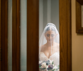 Bride in white dress and veil stands indoors, holding bouquet and looking down in a classic New Orleans wedding scene.