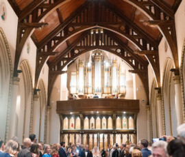 Bride and groom walk down a grand church aisle with wooden arches and organ pipes as guests smile during the ceremony.