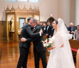 Bride in white dress smiles as two men hug at New Orleans church wedding; guests sit by stained glass windows.