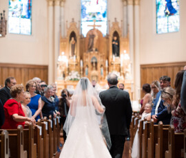 Bride in white gown and veil walks with older man down church aisle, guests seated, stained glass and ornate altar ahead.