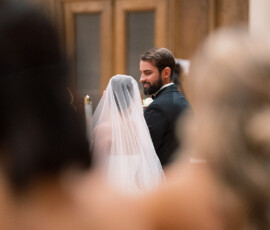Bride in veil and groom stand together indoors, with blurred guests and wooden doors in the background.