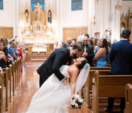 Newlyweds share a romantic kiss as the groom dips the bride, surrounded by guests at their wedding ceremony.