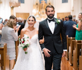 Bride in white gown and groom in black tuxedo smile, walking arm-in-arm down church aisle as guests take photos.