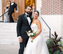 Bride and groom smile outside a building as bridesmaid and groomsman celebrate behind; flowers decorate the charming scene.