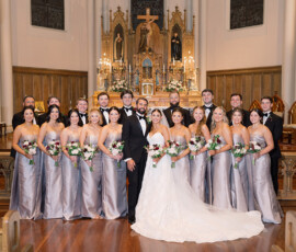Bride and groom with wedding party at church altar, bridesmaids in silver, groomsmen in black suits, all smiling.