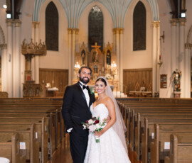 Bride in white gown and groom in black tuxedo stand in ornate church with wooden pews and high arched ceilings.