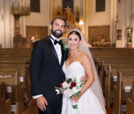 Bride in a white gown and groom in black tuxedo smile inside a church with wooden pews, perfect New Orleans wedding inspiration.