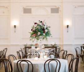 Elegant round table with white cloth, wooden chairs, floral centerpiece, and candles in a bright Louisiana wedding setting.