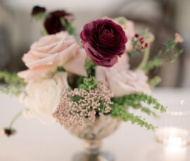 Silver vase with pink, white, and dark purple flowers beside a lit candle on a table; blurred twinkling lights behind.