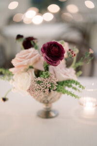Silver vase with pink, white, and dark purple flowers beside a lit candle on a table; blurred twinkling lights behind.
